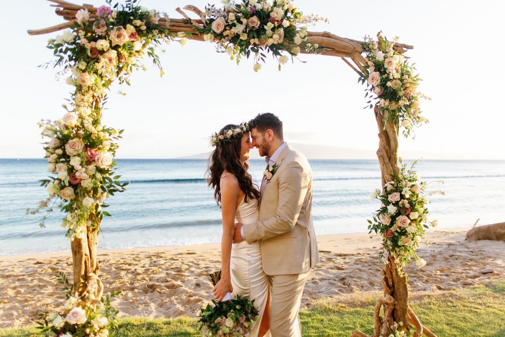 Couple kissing under floral arch on beach wedding ceremony with ocean backdrop and pink roses.