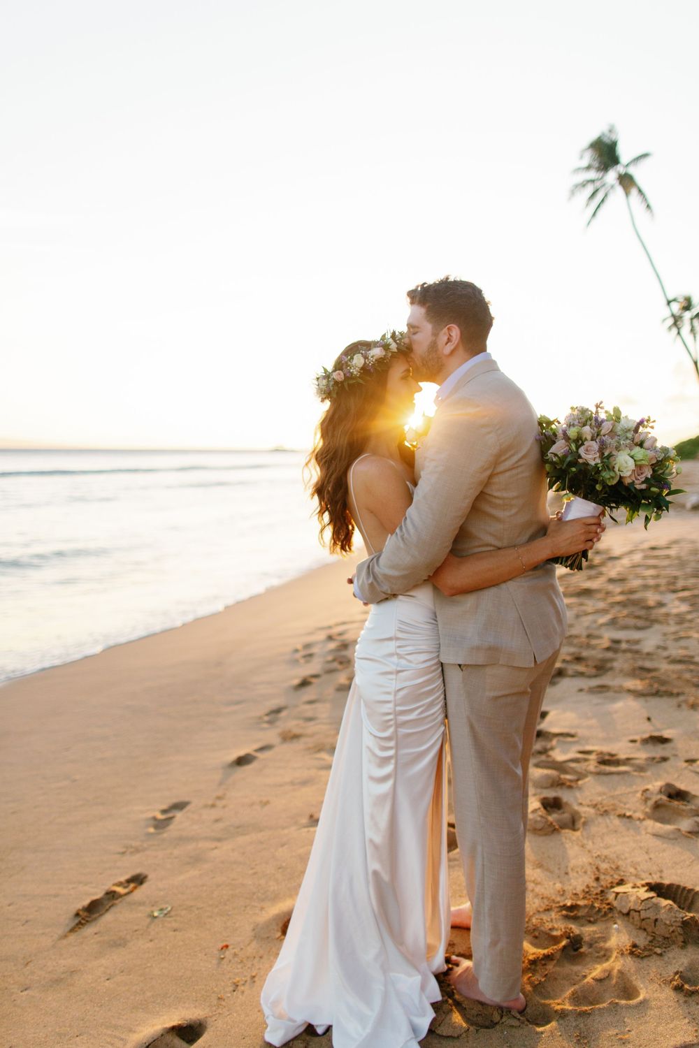 Bride and groom share a romantic sunset kiss on a tropical beach, with palm trees and ocean waves in the background.
