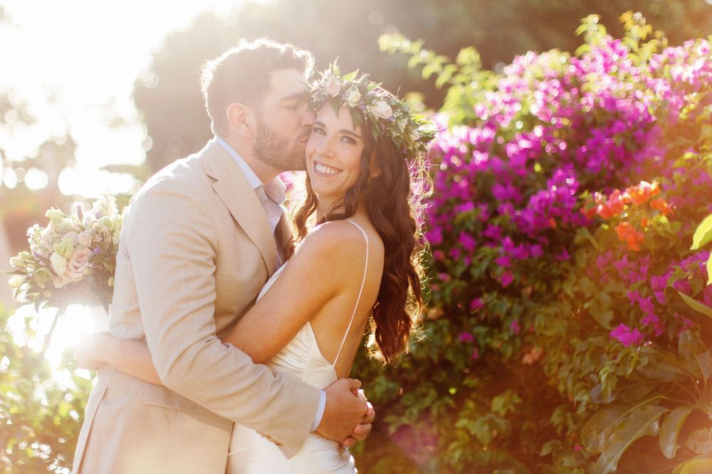Bride and groom embrace outdoors with vibrant pink bougainvillea flowers in sunlit garden wedding photo.