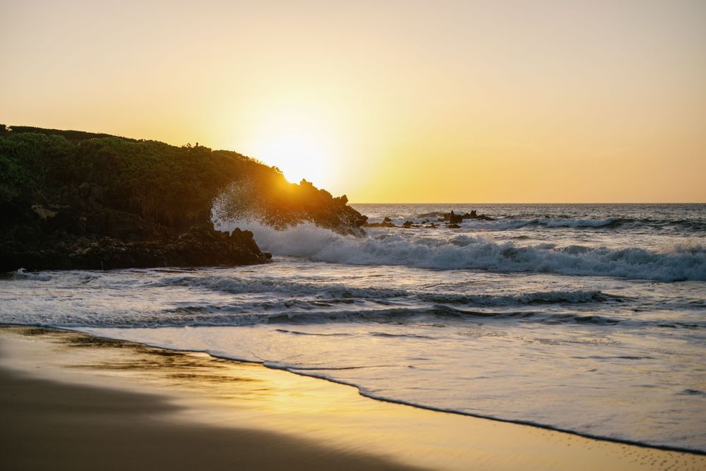Golden sunset over rocky coastline with crashing ocean waves and sandy beach.