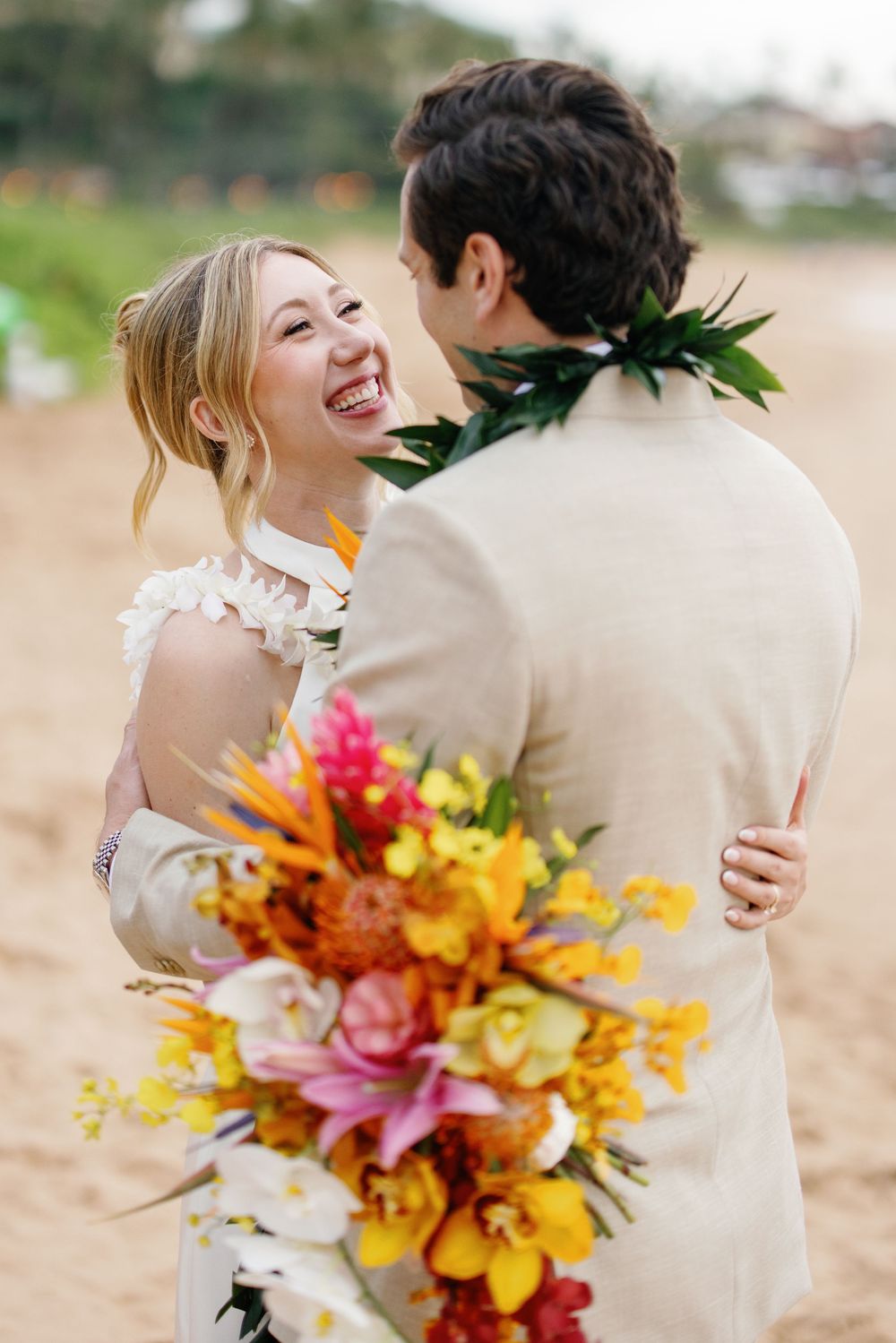 Laughing bride holding tropical bouquet embraces groom on sandy beach during Hawaiian wedding ceremony.