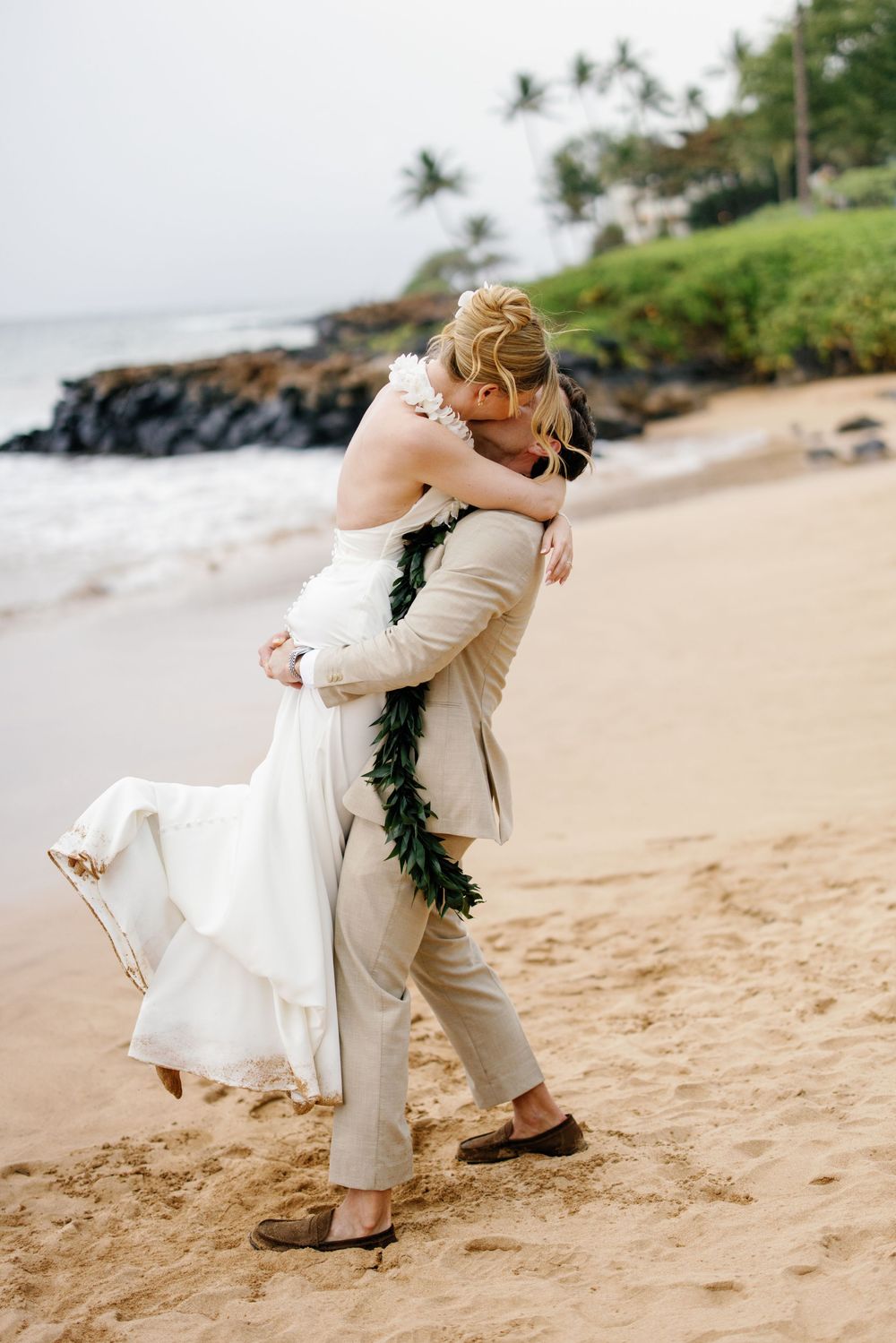 Groom lifting bride in white dress on sandy Hawaii beach, sharing romantic kiss with tropical ocean backdrop.