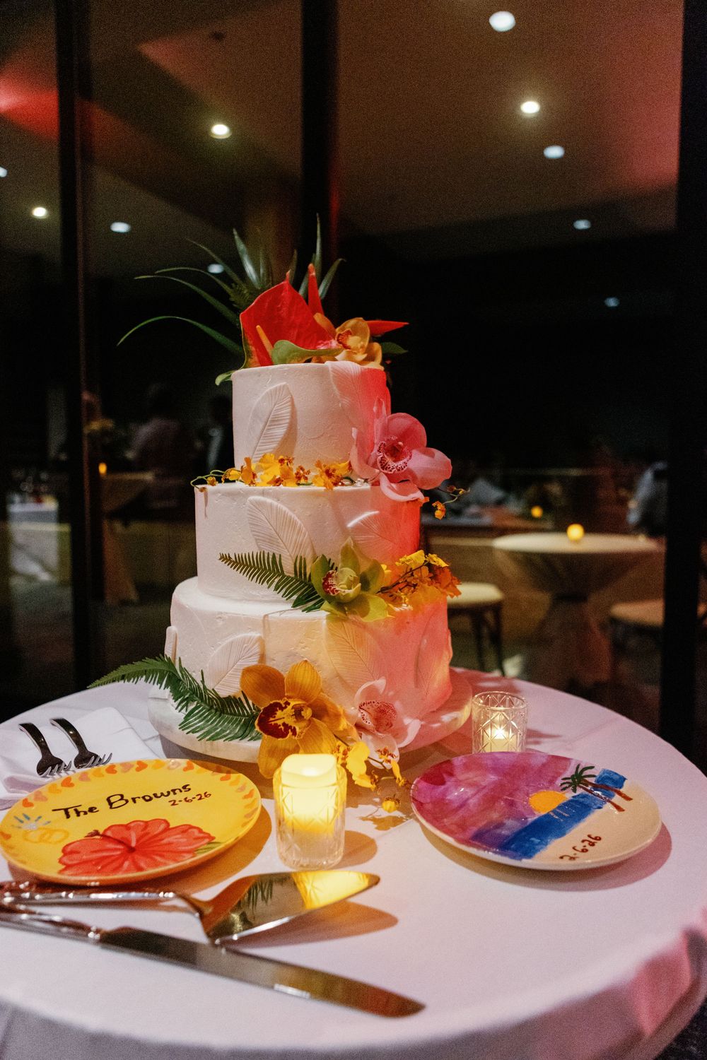 Tropical three-tier white wedding cake with orchids and anthurium flowers on a table with colorful plates and candles.