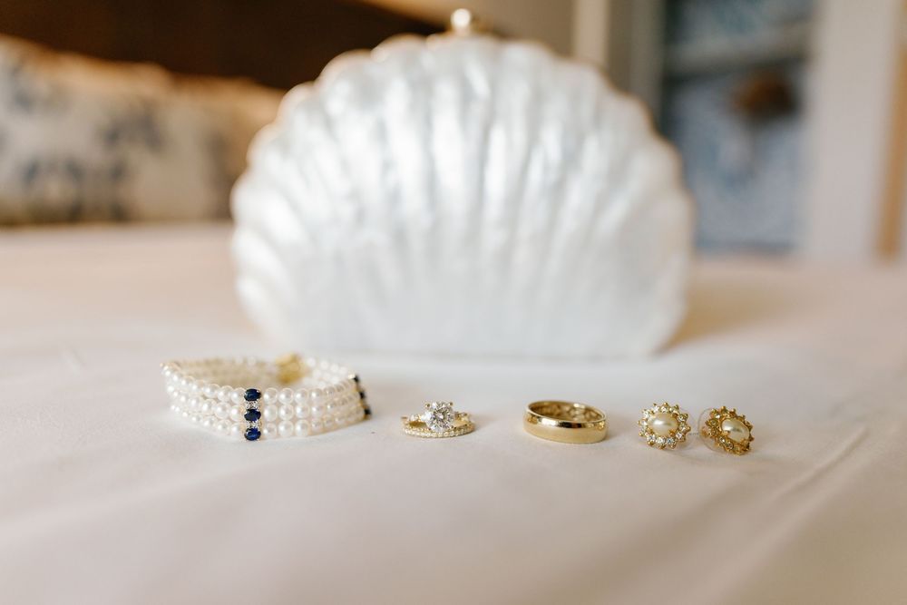 Pearl bracelet, gold rings, and pearl earrings arranged on white fabric beside a shell-shaped clutch purse.