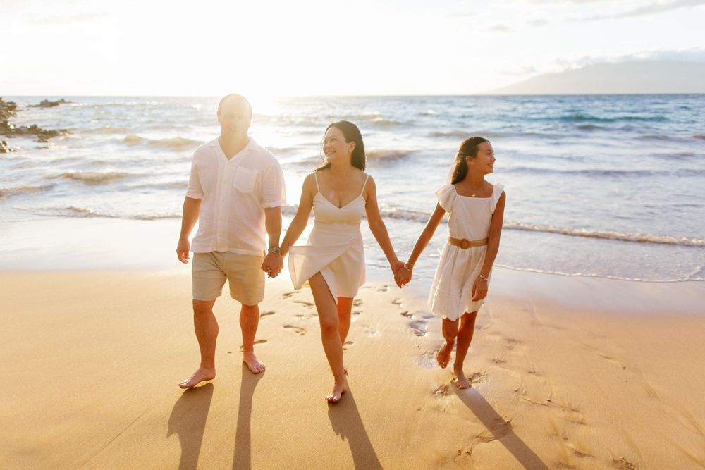 Family of three walking hand-in-hand on a sunny beach at golden hour, wearing white outfits near ocean waves.