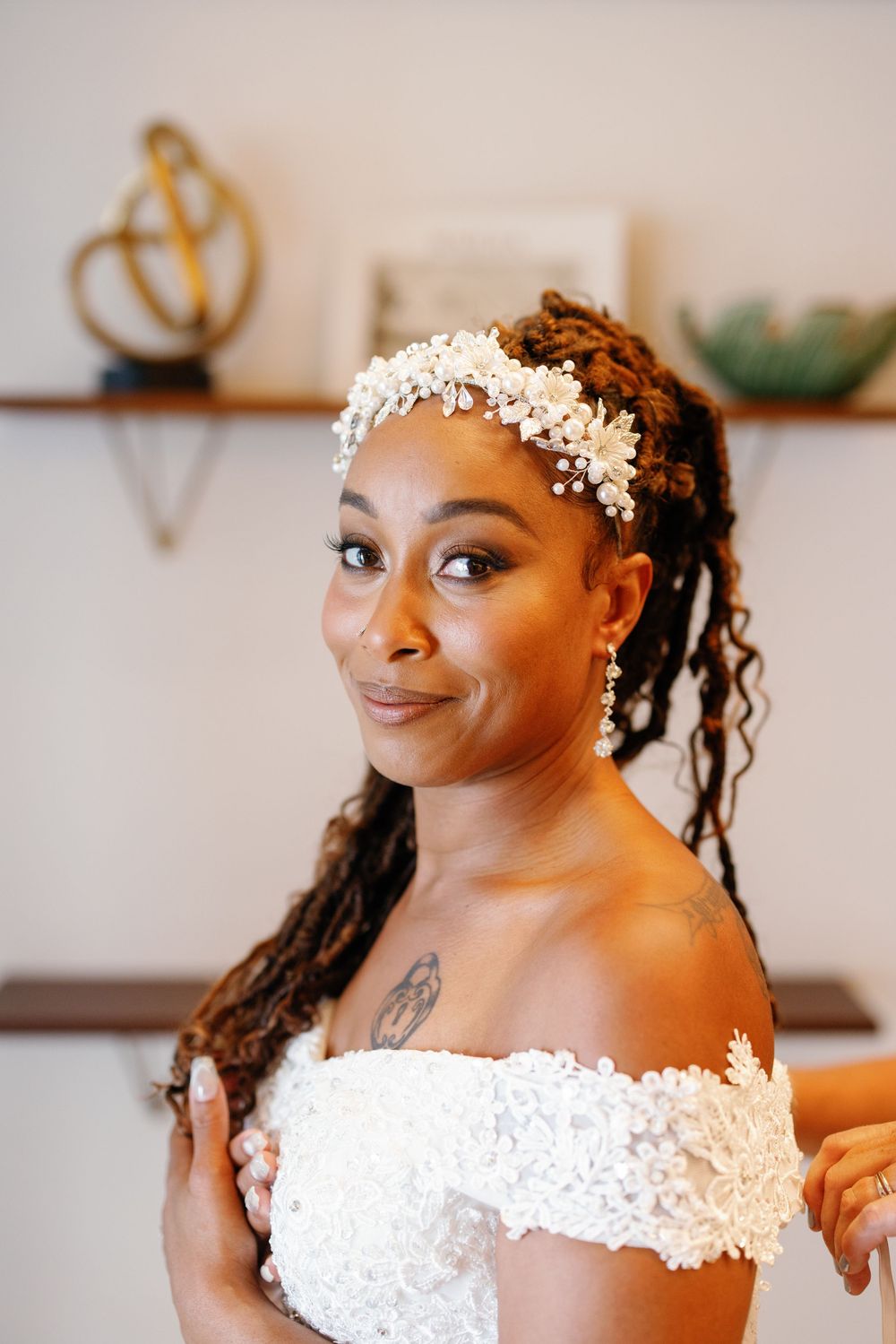 A Black bride in a white off-shoulder lace gown and floral headpiece smiles warmly, showcasing elegant drop earrings and a tattoo.