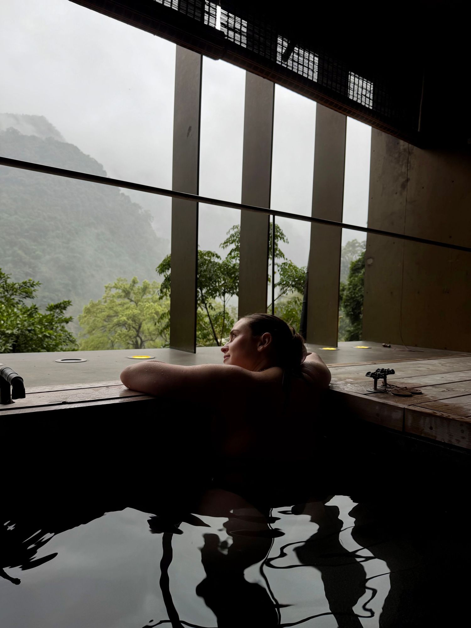 Person relaxing in an indoor hot spring bath with misty mountain views through large windows.