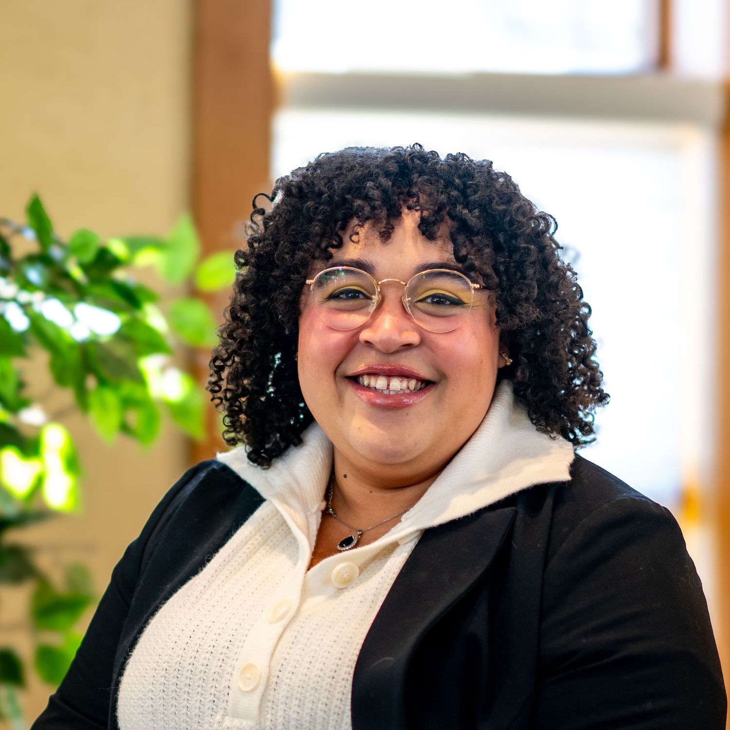 Smiling woman with curly hair and glasses wearing a black blazer, posed in a bright modern office setting.