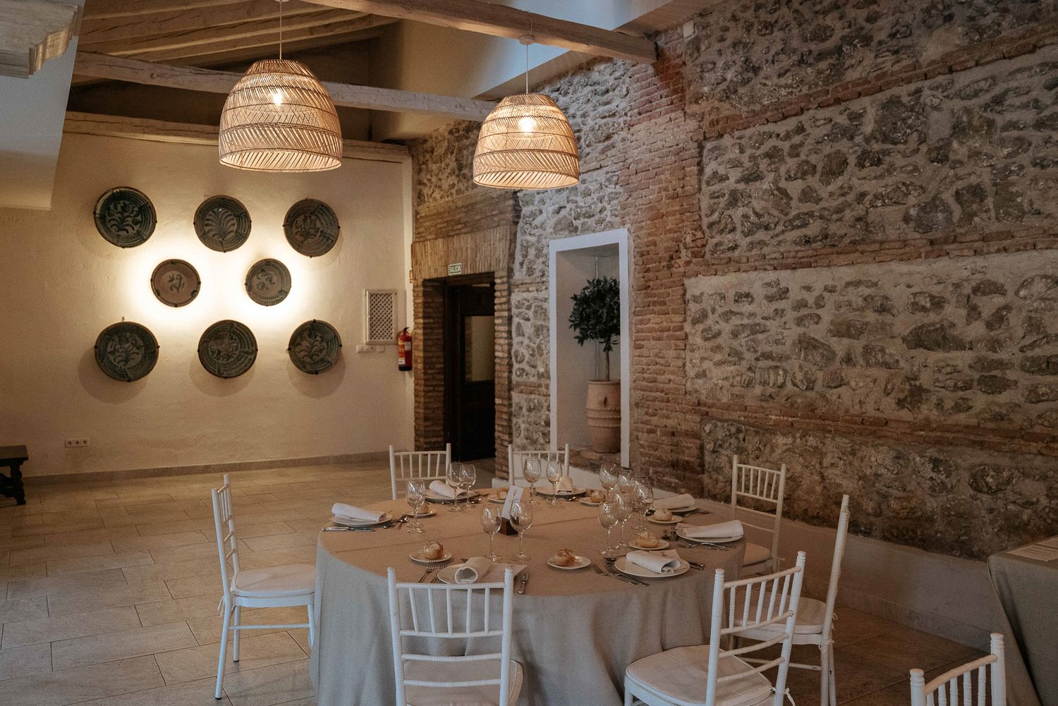 Elegant rustic dining room with stone walls, wicker pendant lights, round tables with white chairs, and decorative plates on display.
