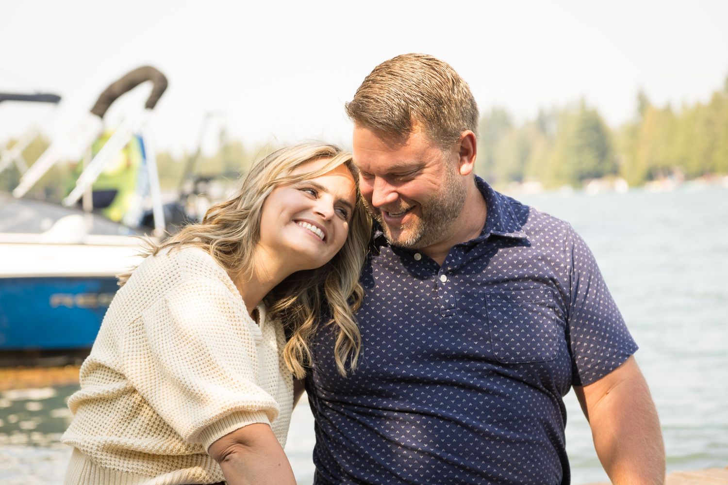 Happy couple smiling and embracing near a lake with a boat in the background on a sunny day.
