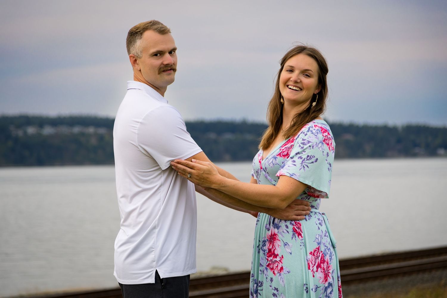 Couple holding hands and smiling near waterfront with trees and cloudy sky in background.