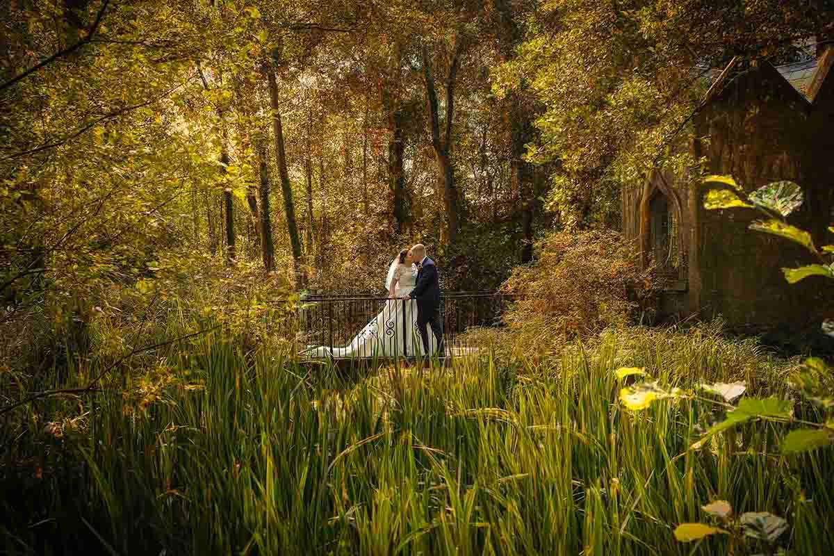 A bride and groom embrace in a lush green forest garden setting with golden autumn foliage and tall grass.