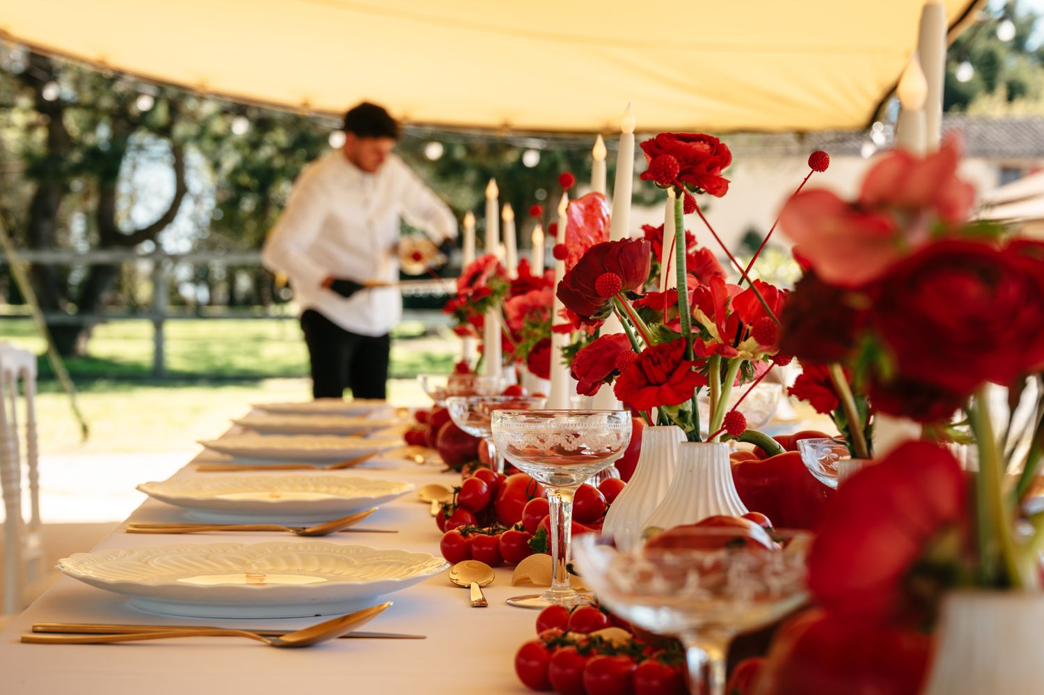Long decorated banquet table with red roses, white candles, and gold cutlery under a tent.