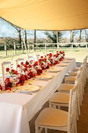 Une table de réception de mariage en plein air, ornée de compositions florales rouges et de chaises blanches, sous un auvent.