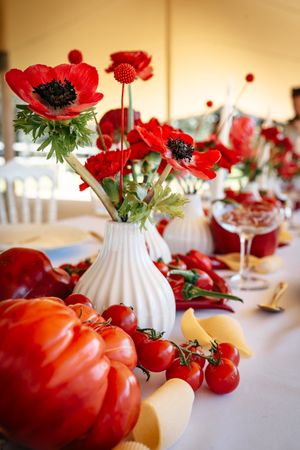 White vases with red anemone flowers surrounded by tomatoes and peppers on a wedding table.