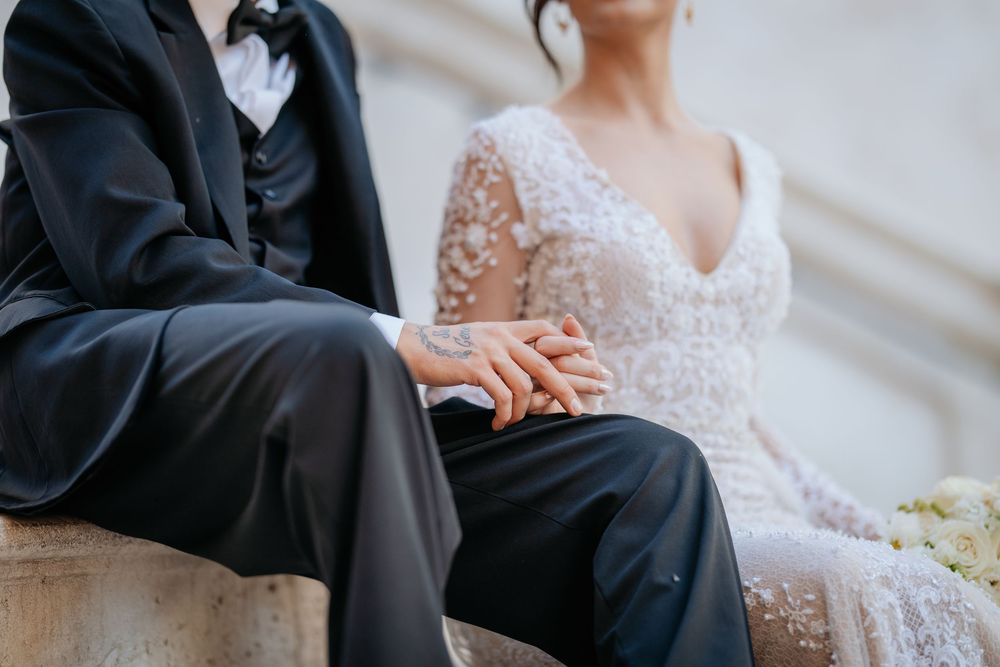 Bride and groom sitting together holding hands in elegant wedding attire outdoors.