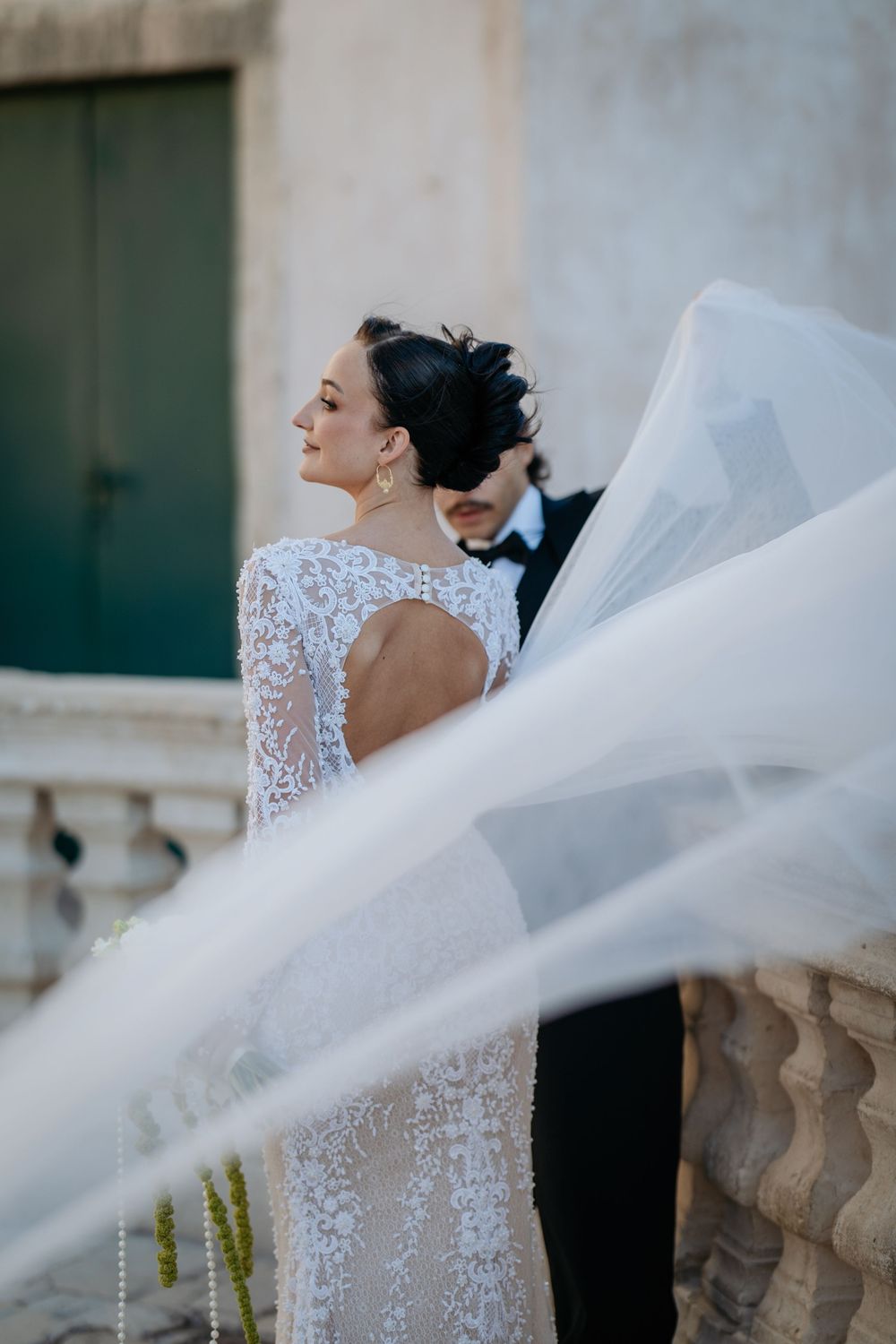 Bride in backless lace gown with flowing veil billowing in the wind outdoors.