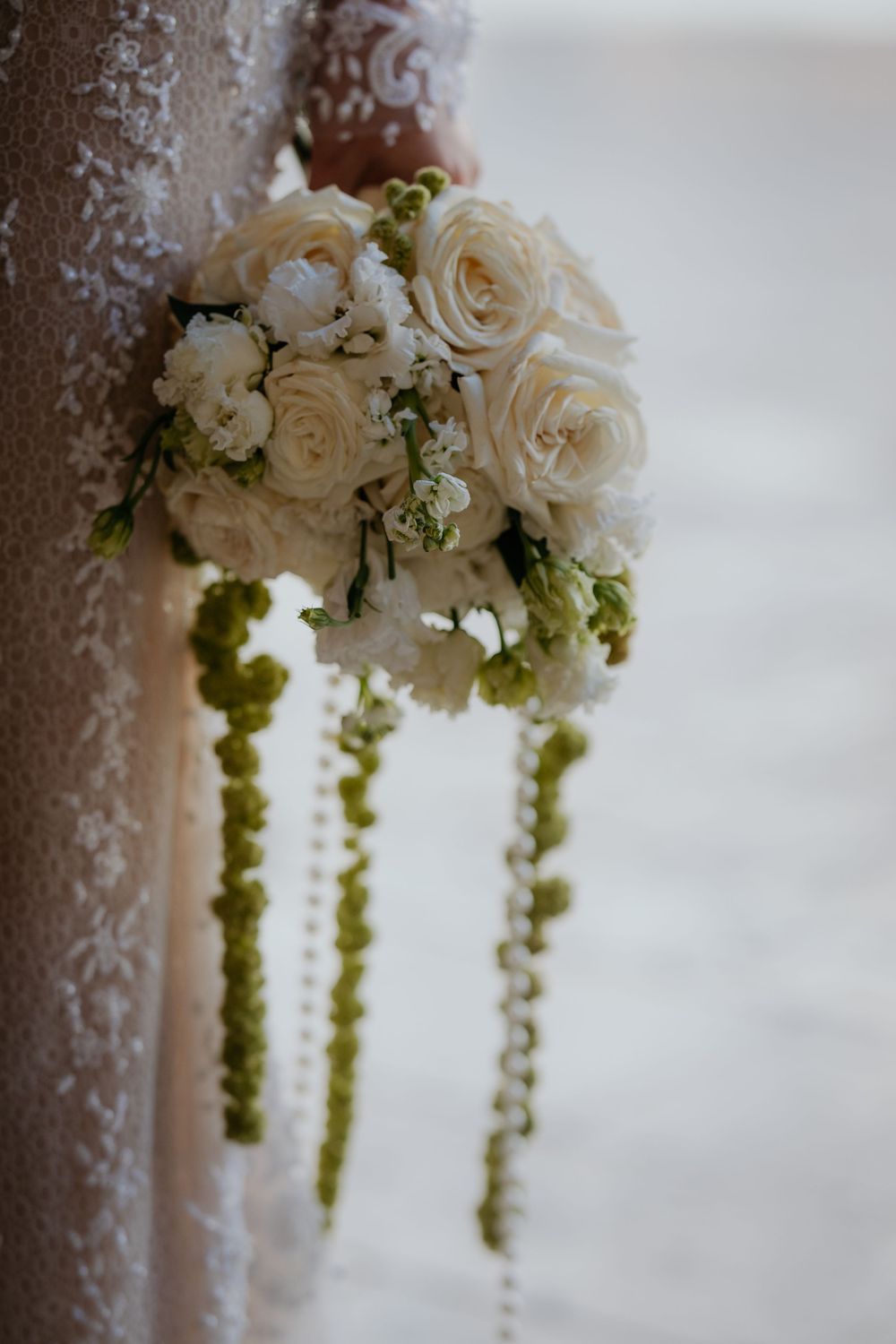 Bride holding elegant white rose bouquet with cascading green pearl strands in lace dress.