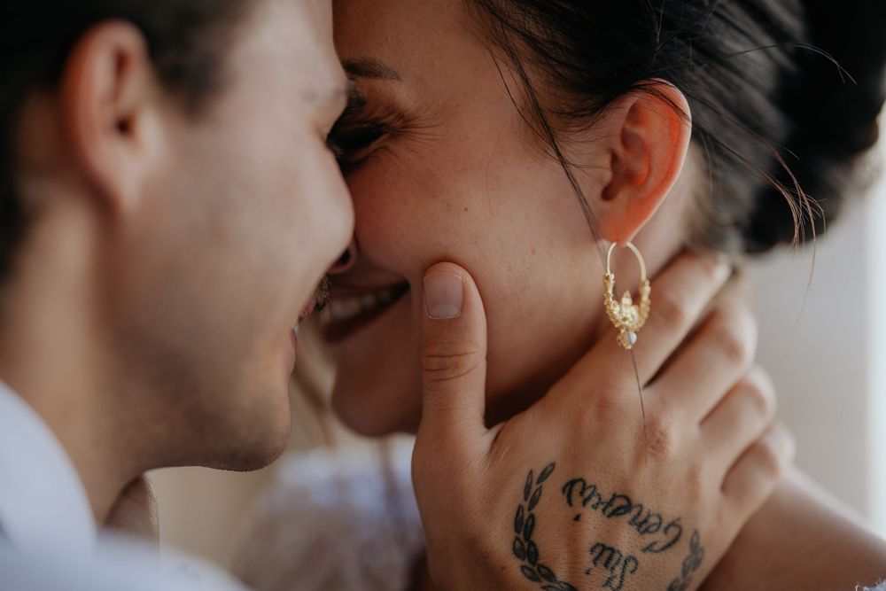 Couple sharing intimate moment, bride smiling with gold hoop earring and tattooed hand visible.