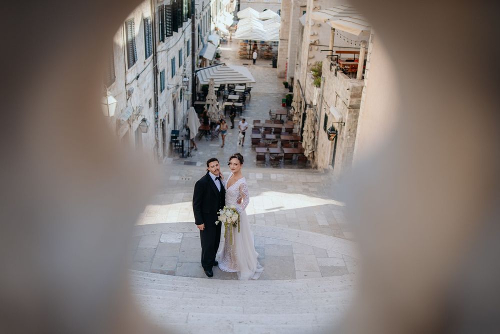 Bride and groom posing on historic stone steps framed by circular architectural opening in Dubrovnik.