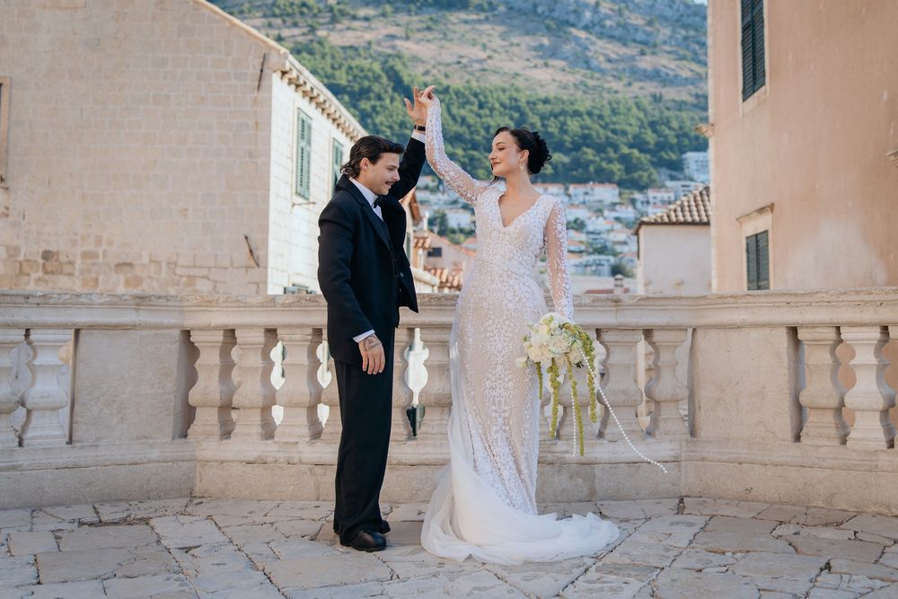 Joyful bride and groom celebrating on terrace balustrade with scenic Dubrovnik hillside backdrop.