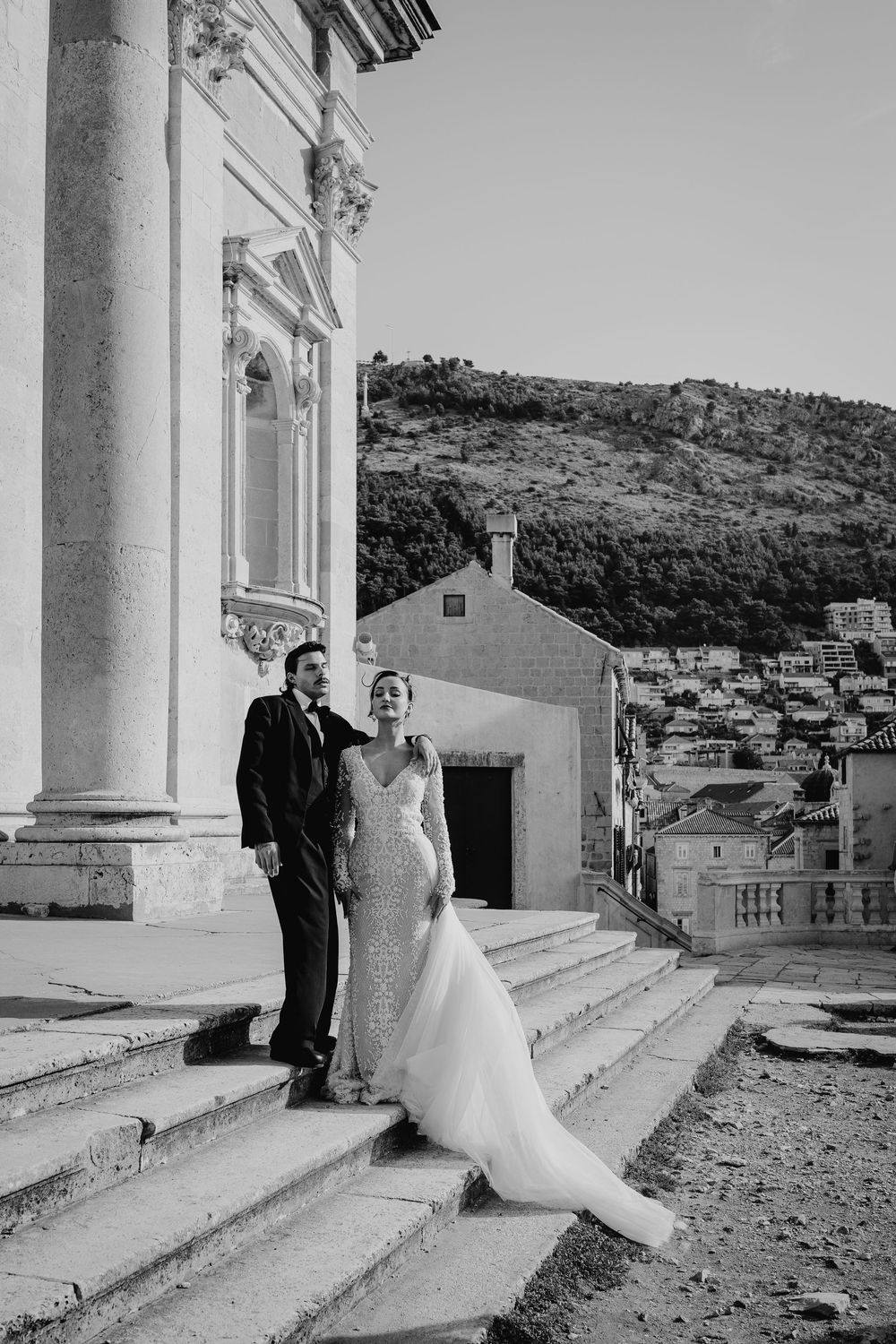 Black and white photo of bride and groom posing on stone steps beside a classical European church building.