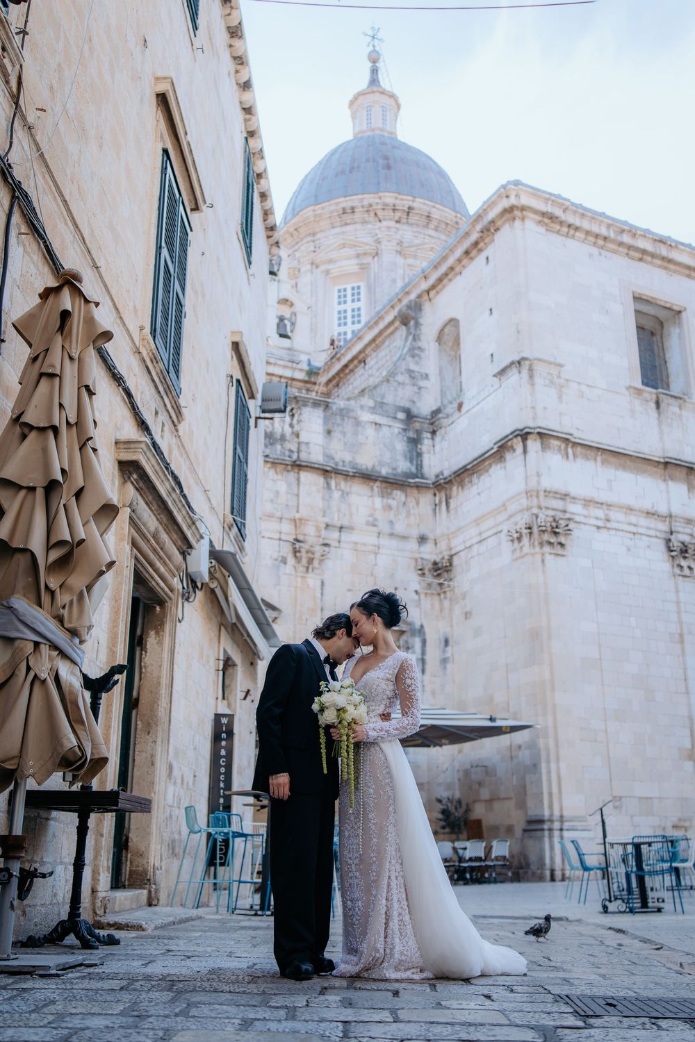 Wedding couple sharing a romantic kiss in a cobblestone square with a historic domed cathedral backdrop.