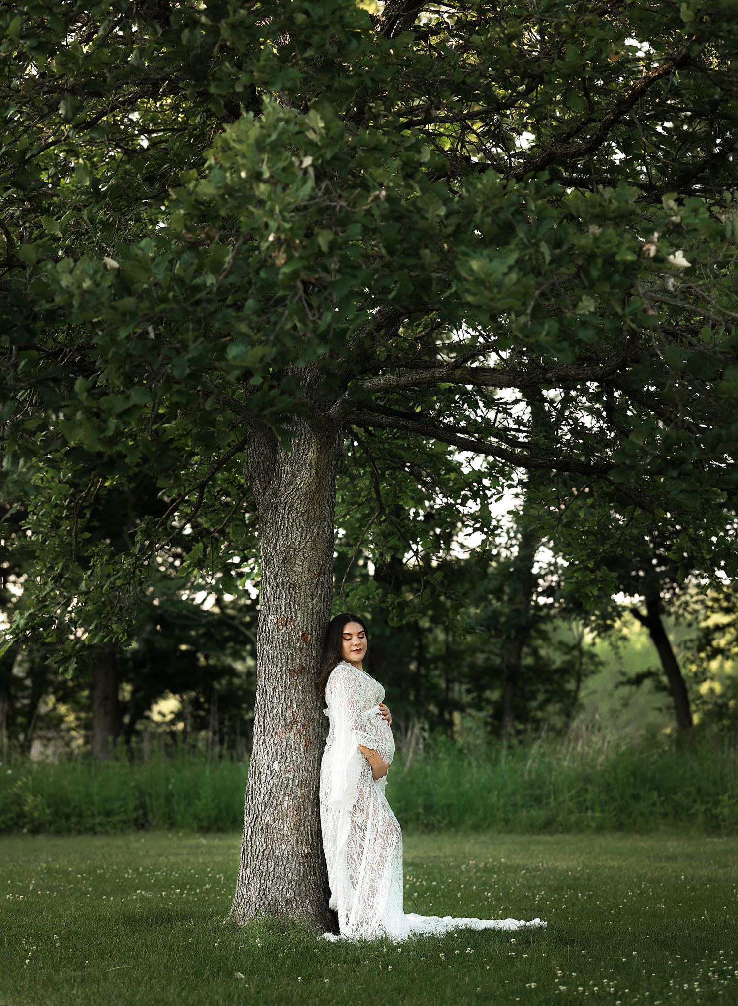 Pregnant woman in white lace gown leaning against a large tree in a lush green park during a maternity photo session.