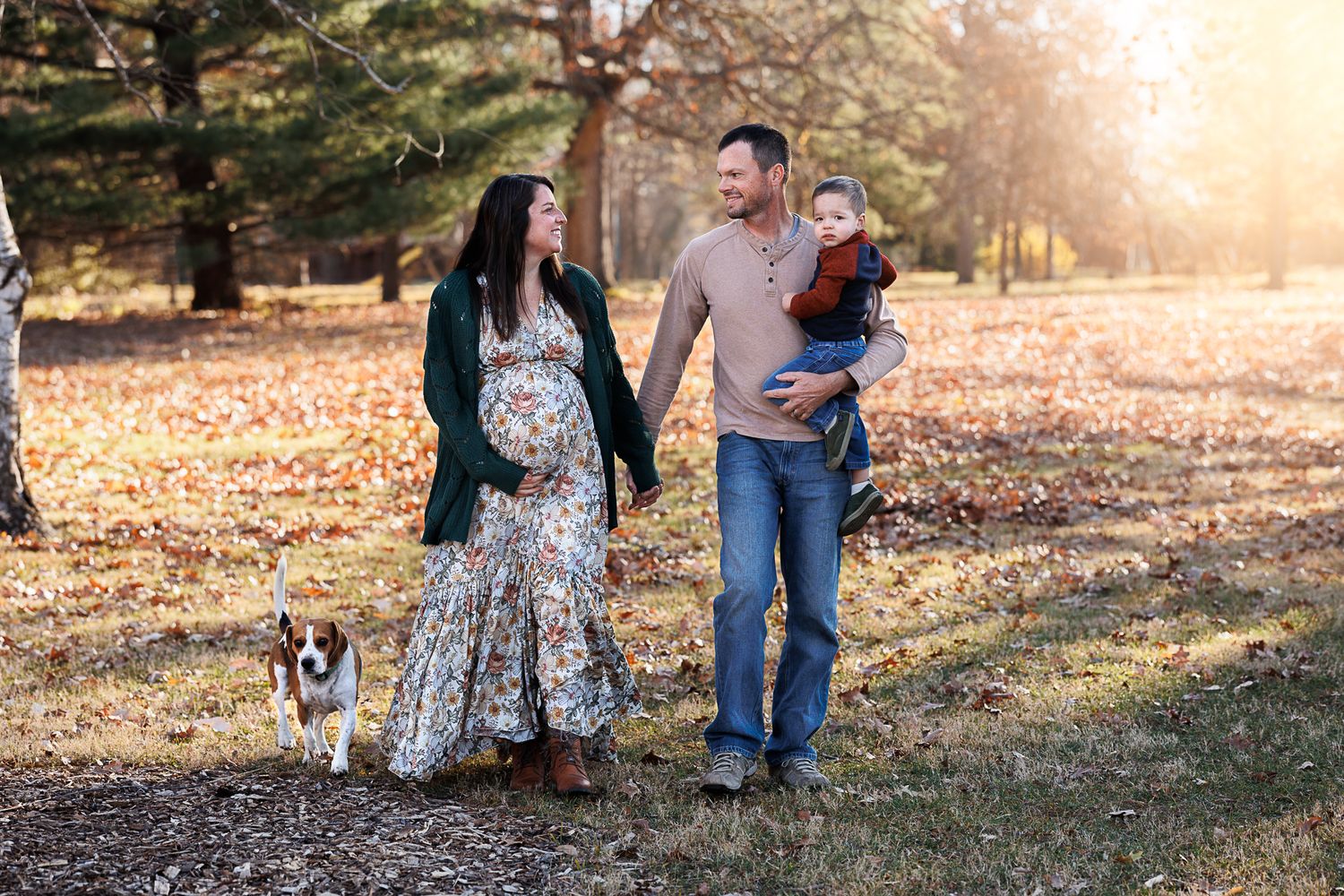 Pregnant woman in floral dress walks with husband holding toddler and dog in sunlit autumn park.