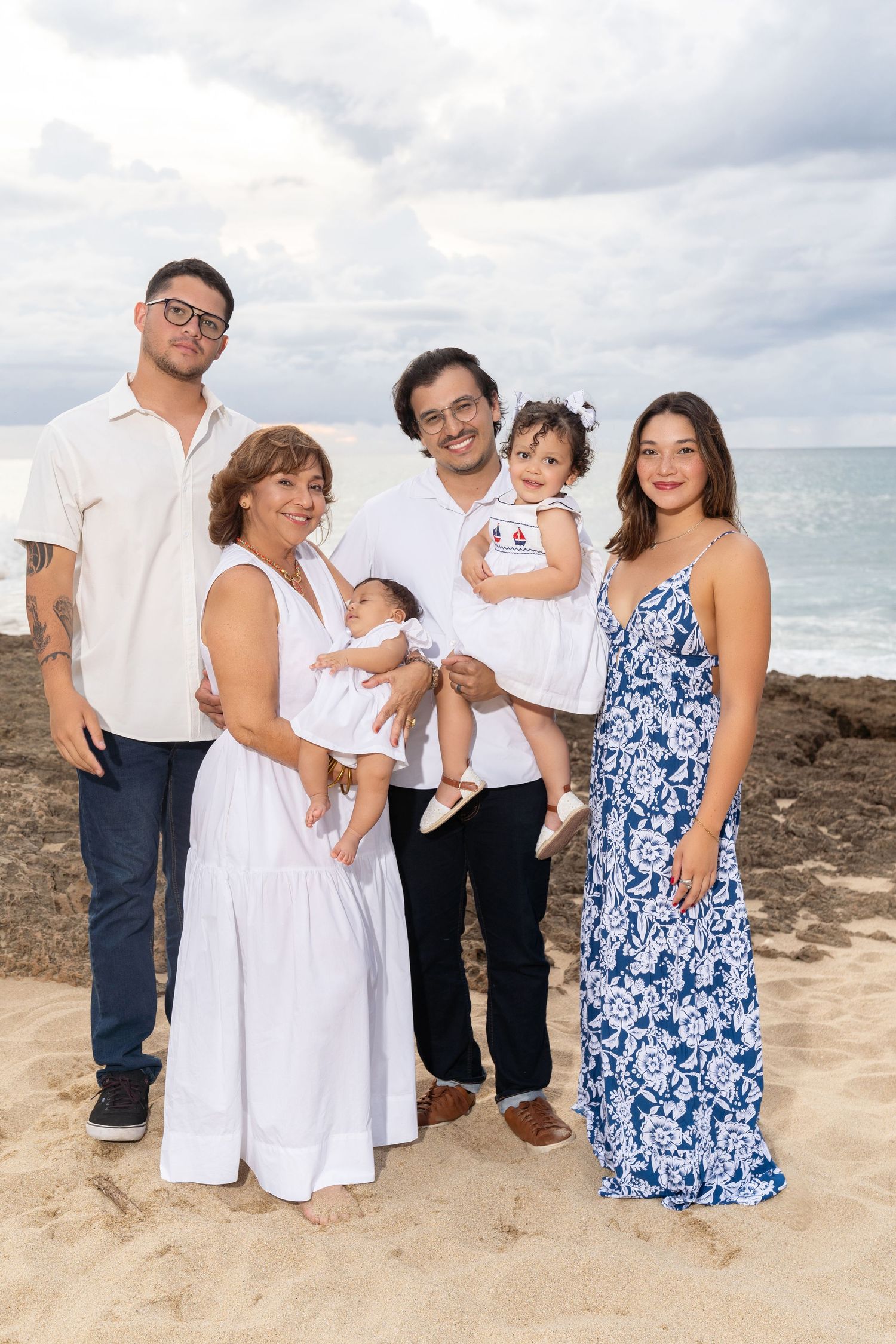 Four people smiling together on a rocky beach with ocean waves and cloudy skies in the background.