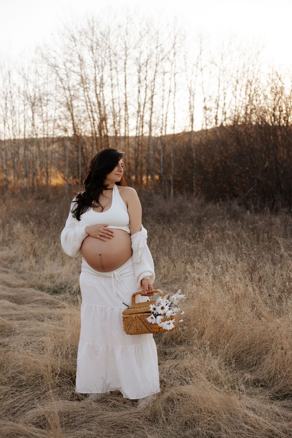 Pregnant woman in white outfit holding a wicker basket with flowers in a golden field at sunset.