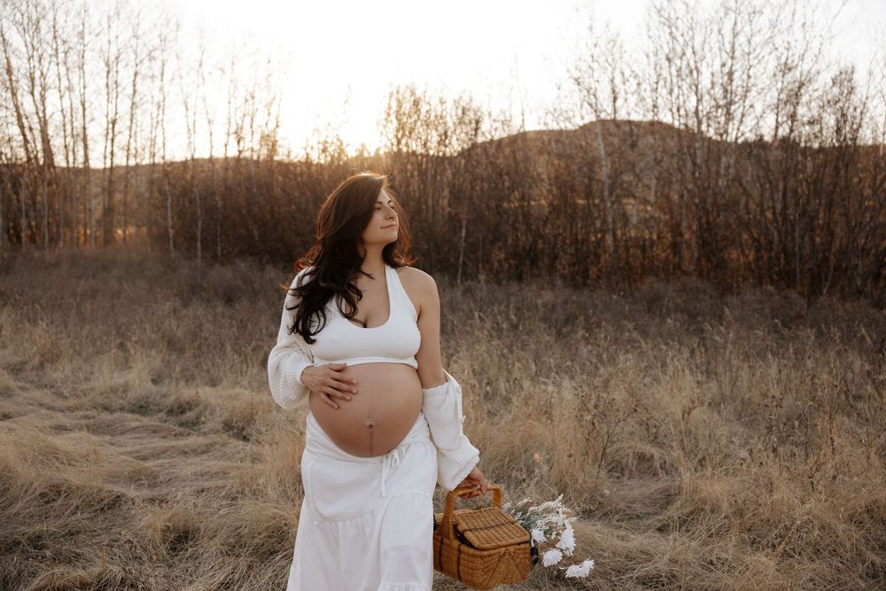 Pregnant woman in white dress holding baby bump in golden field at sunset with wicker basket and wildflowers.