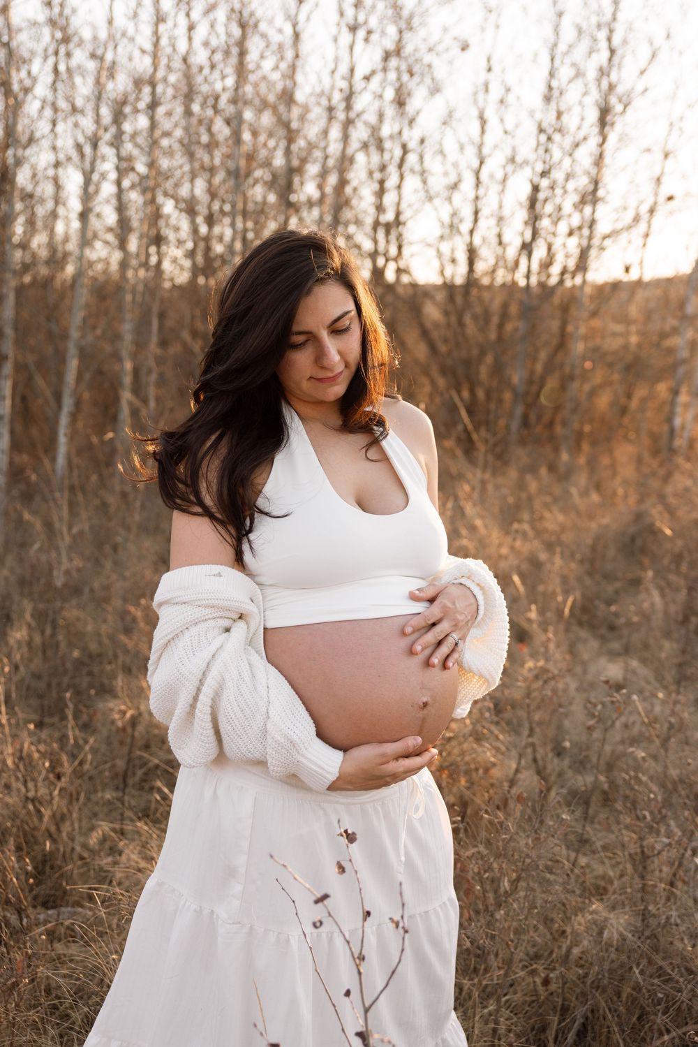 Pregnant woman in white crop top and skirt cradling baby bump outdoors at golden hour.