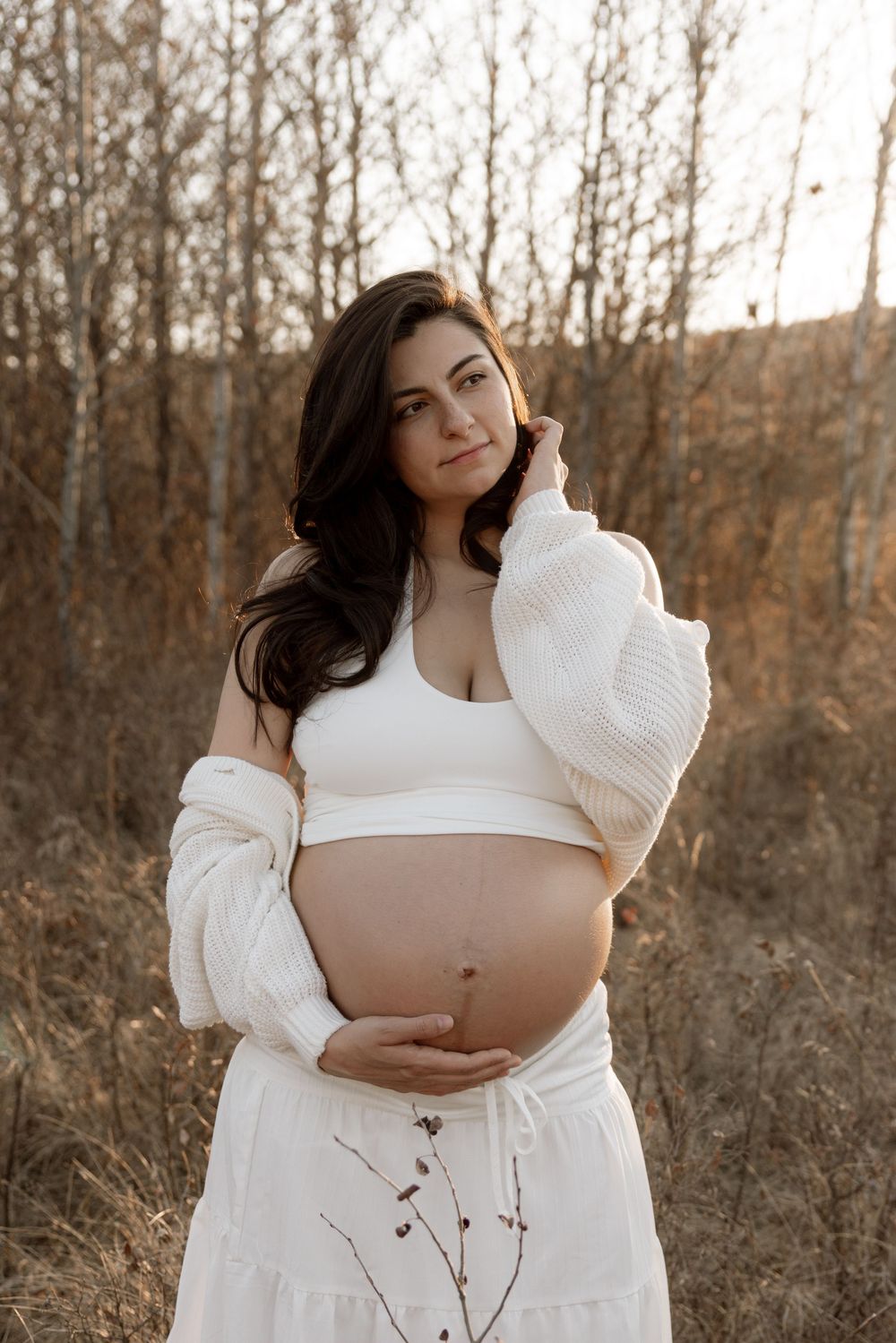 Pregnant woman in white crop top and skirt cradling baby bump outdoors in golden hour light.