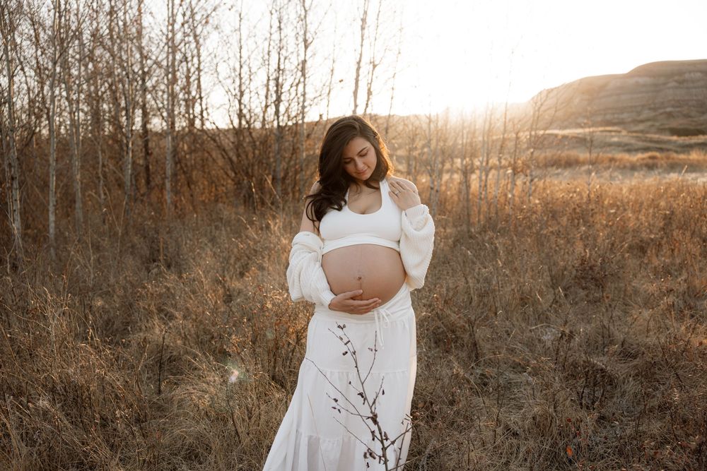 Smiling pregnant woman in white maternity outfit standing in sunlit autumn in calgary alberta for a maternity photoshoot