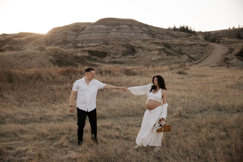 Pregnant couple holding hands in golden field with badlands hills at sunset, in calgary
