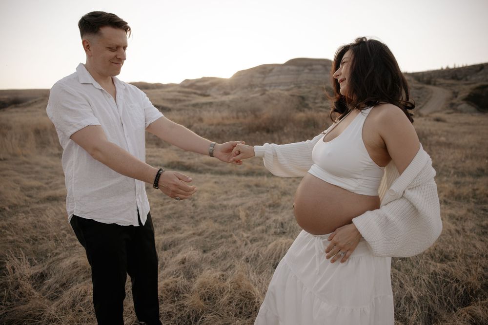 Man reaches out to hold hands with pregnant woman in white dress during outdoor maternity shoot.