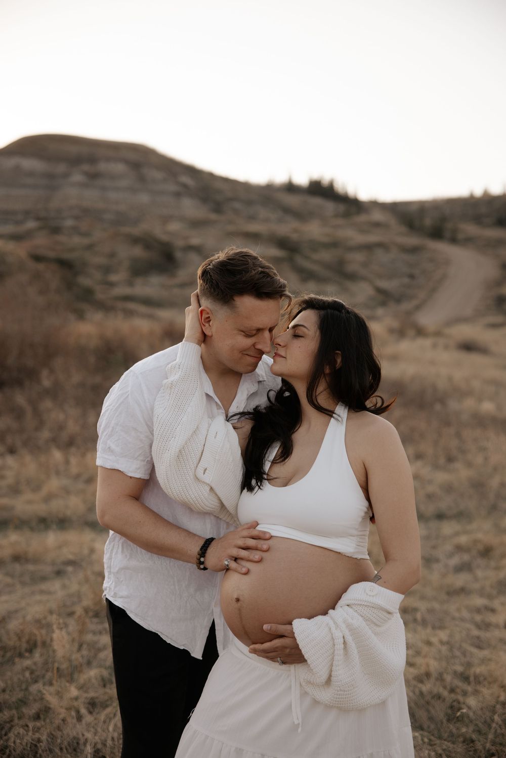 Couple sharing a tender moment outdoors, pregnant woman in white dress cradling baby bump, man embracing her in golden field.
