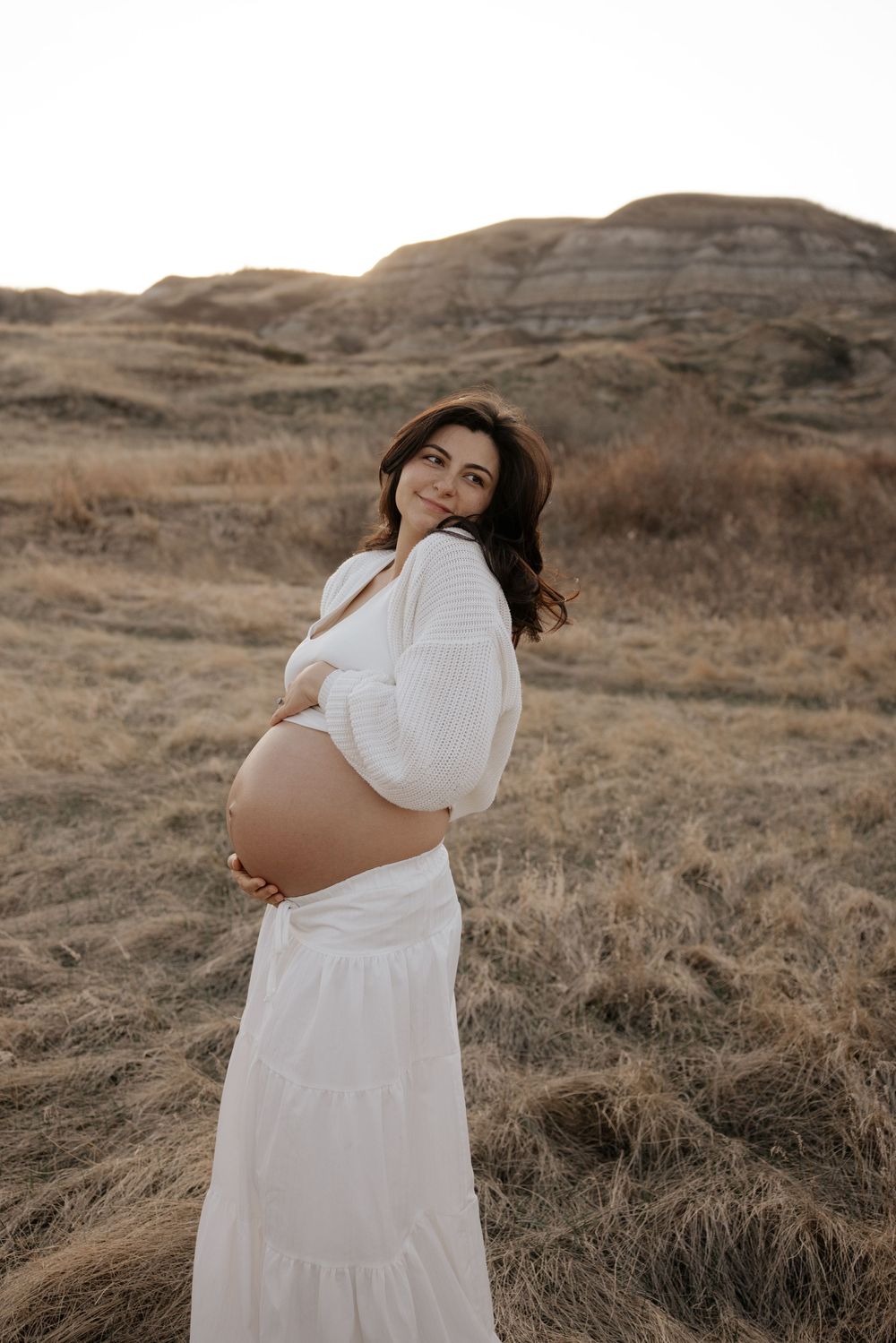 Smiling pregnant woman in white dress cradling her baby bump in a golden field with rolling hills at sunset.