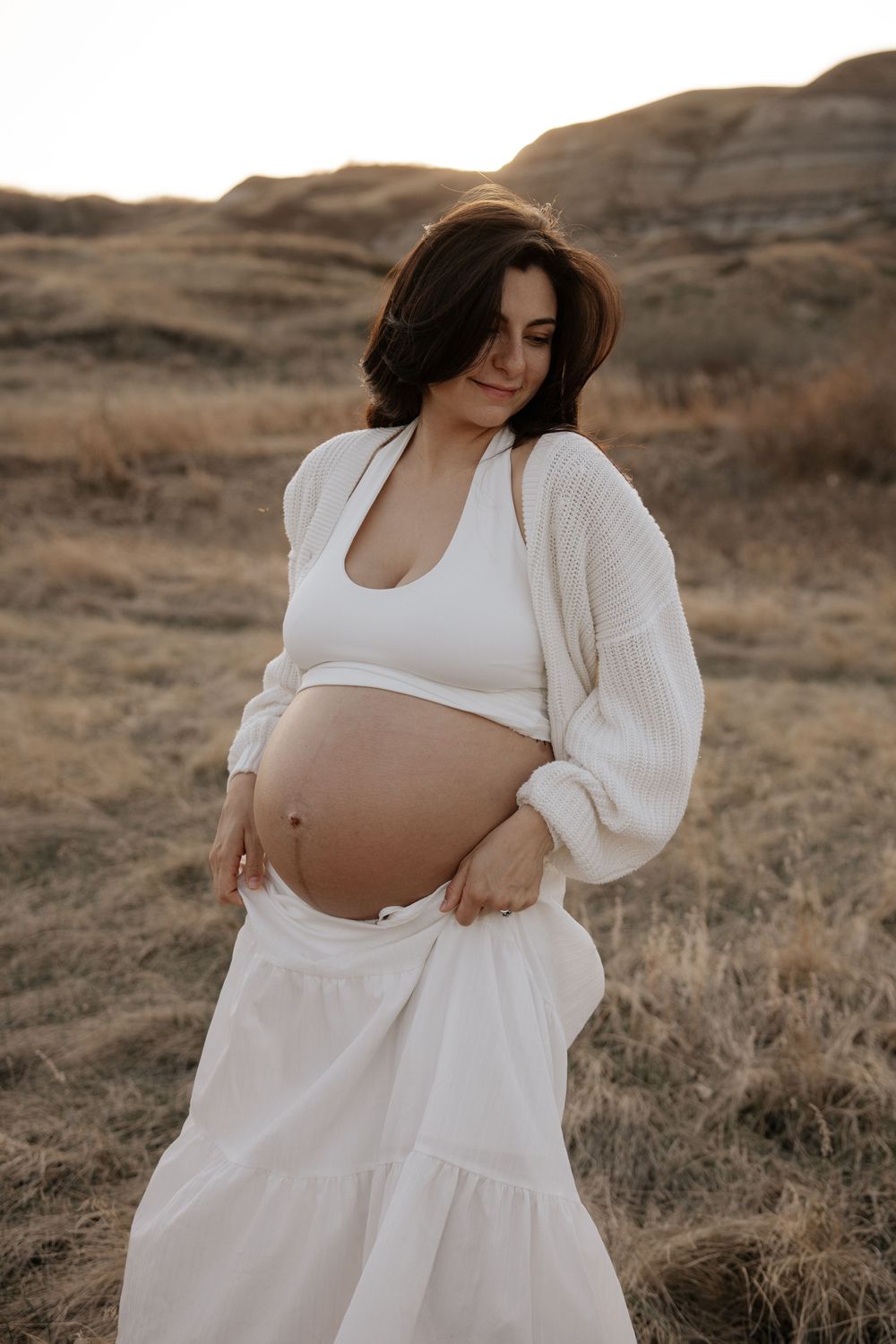 Pregnant woman in white ensemble gently holding baby bump in open field during warm sunset.