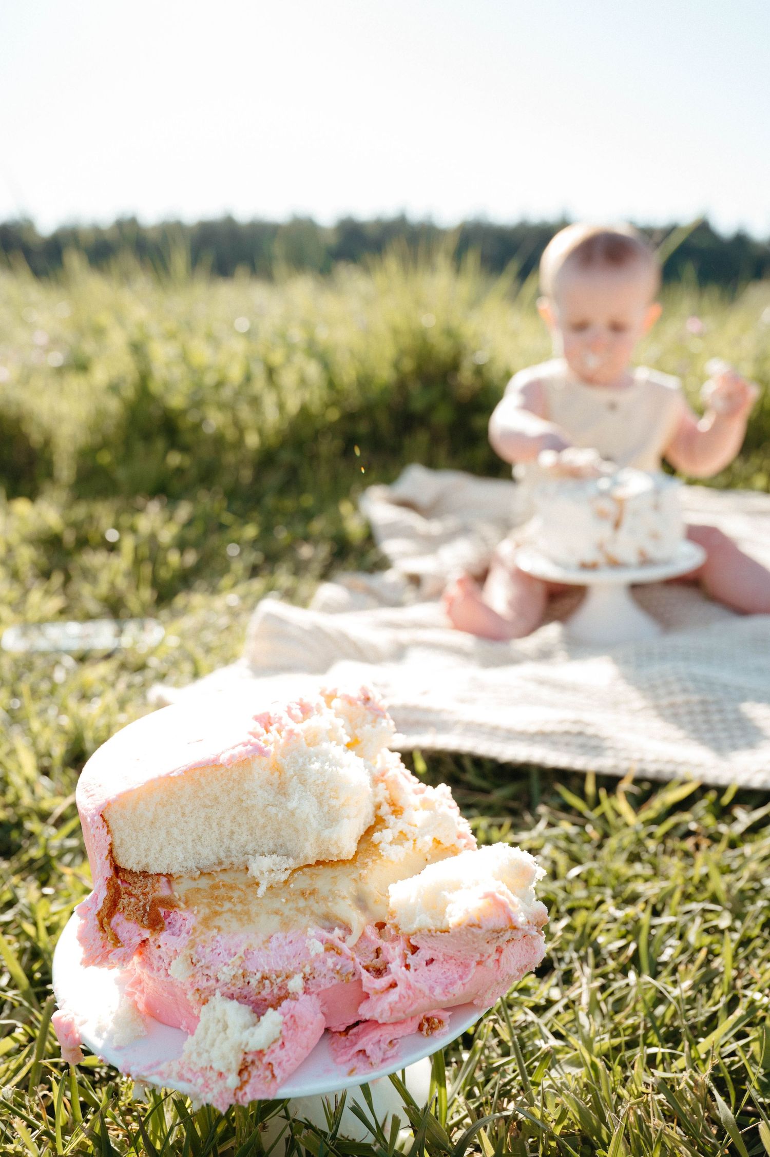 Baby enjoying first birthday cake smash outdoors on grass, with pink and white frosted cake in foreground.