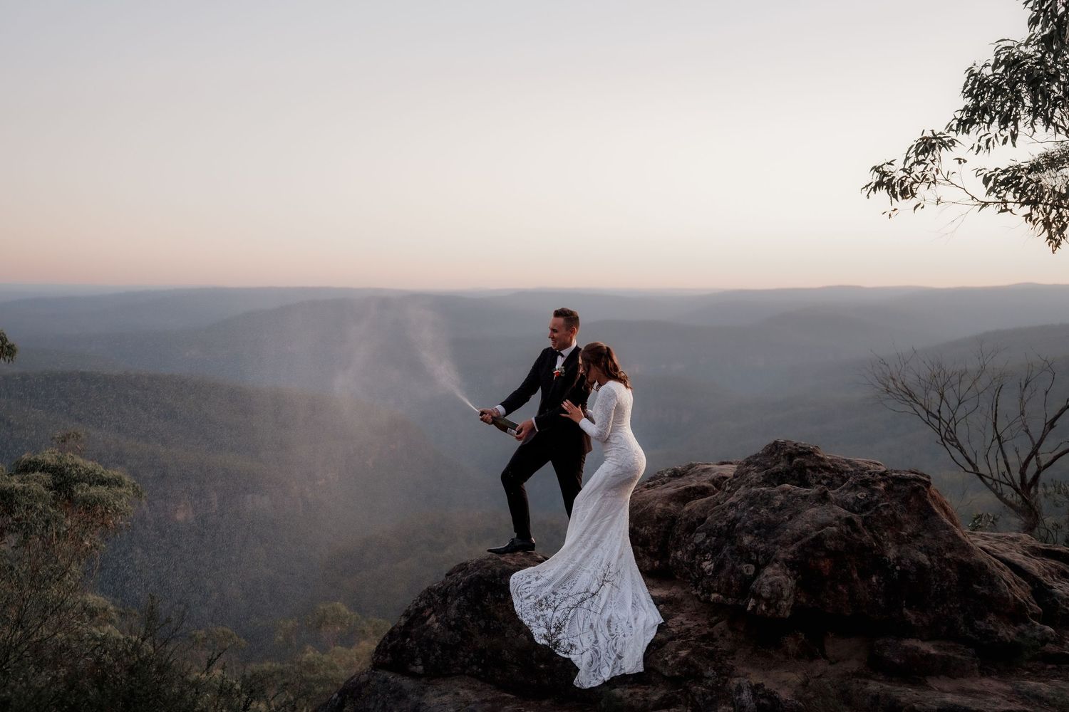Bride and groom stand on a rocky cliff at sunset, overlooking a vast mountain valley landscape.