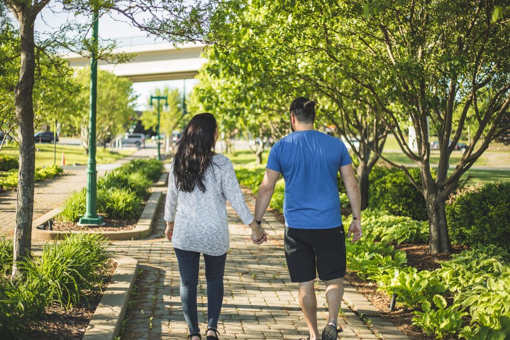 Couple holding hands walking along a lush green tree-lined brick pathway in a sunny park.