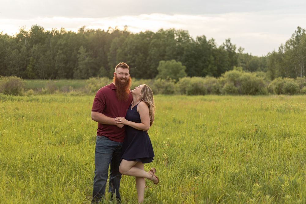 Couple laughing together in a lush green field at golden hour, man with red beard in maroon shirt, woman in navy dress.