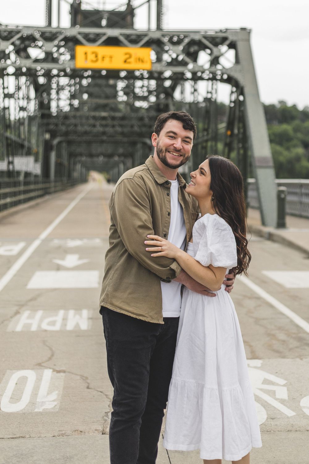 Happy couple embracing on a bridge with a 13ft 2in clearance sign, man in olive shirt and woman in white dress.