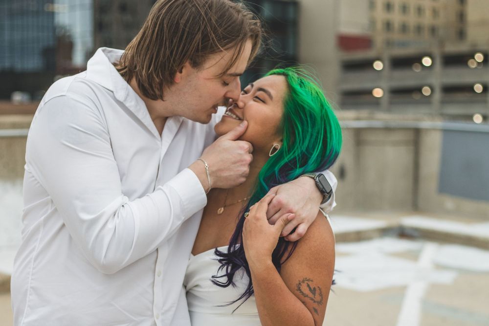 Couple sharing a romantic moment, woman with vibrant green hair embracing man in white shirt at outdoor urban wedding.