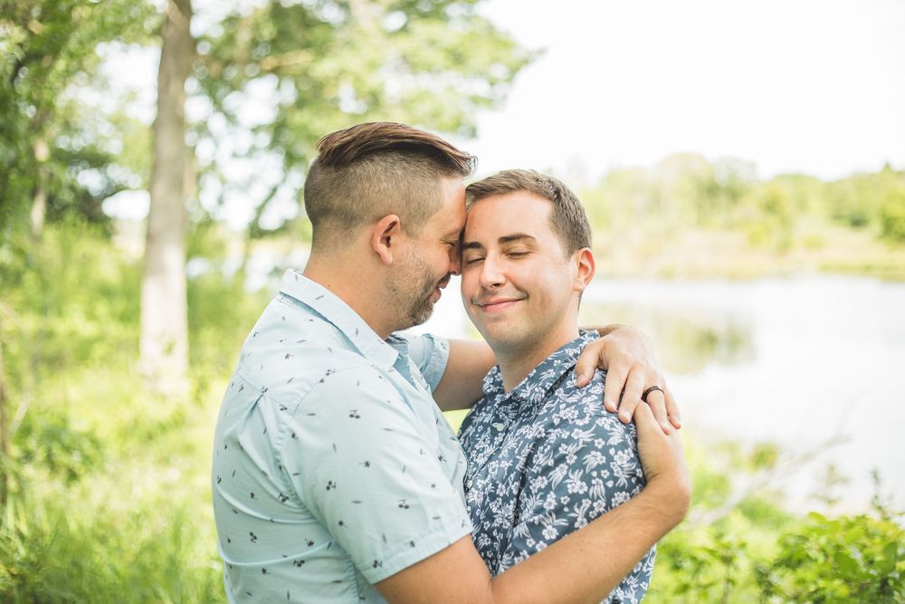 Two men sharing a tender embrace outdoors near a lake, surrounded by lush green trees on a sunny day.