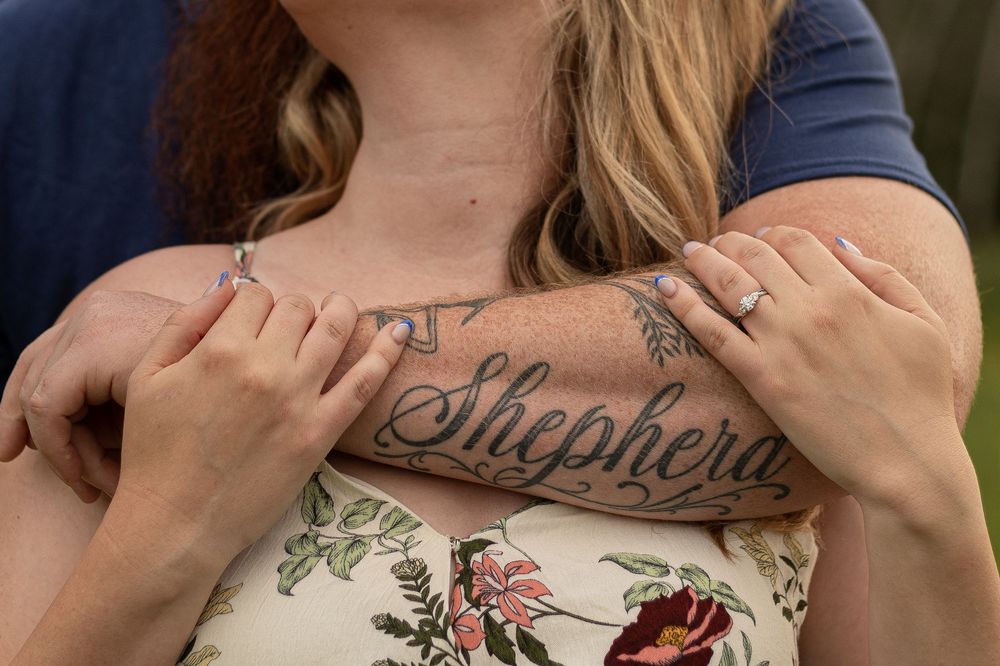 Close-up of a woman with a Shepherdson chest tattoo being embraced by a man, both wearing rings.