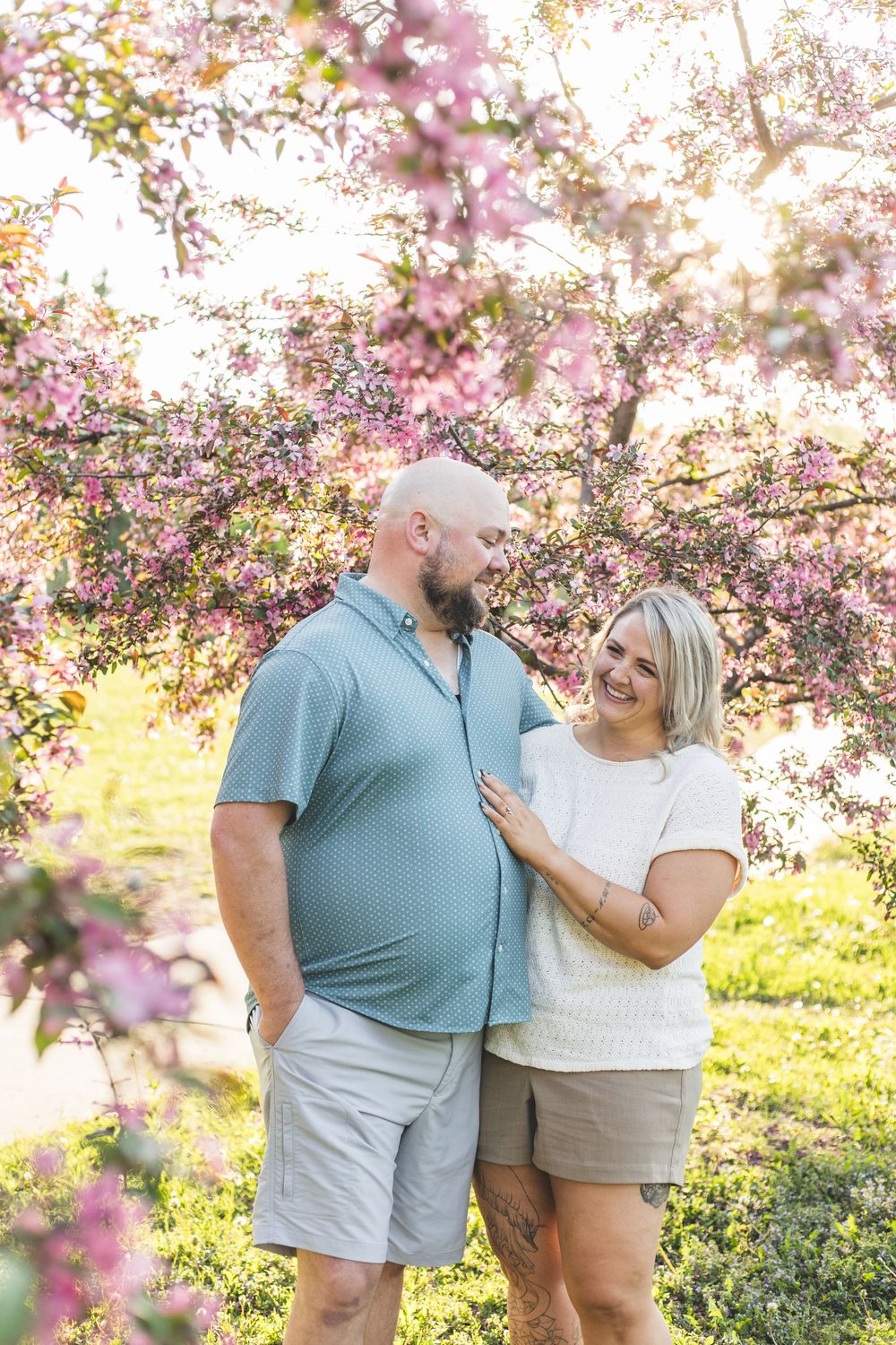 A happy couple embraces and smiles at each other under blooming pink cherry blossom trees in golden sunlight.