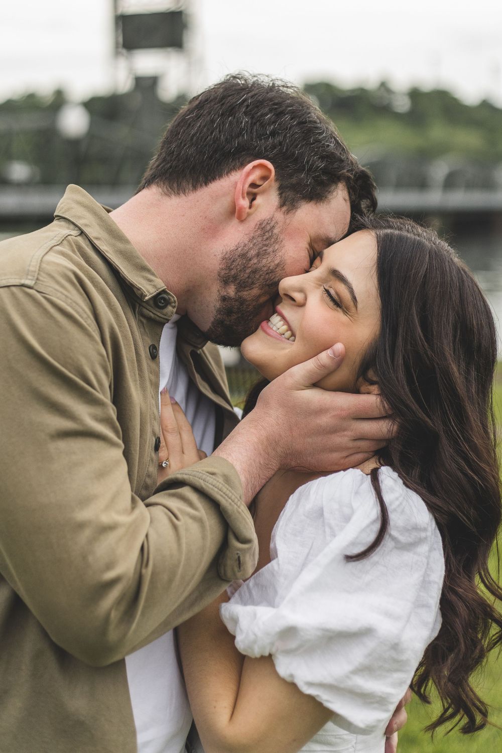 A bearded man in olive jacket tenderly kisses smiling brunette woman's cheek outdoors.